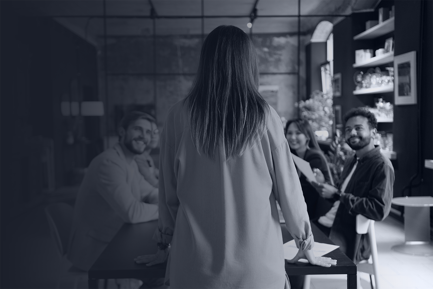 A monotone dark blue photo of a woman to a group in a board room to represent information security policy coaching